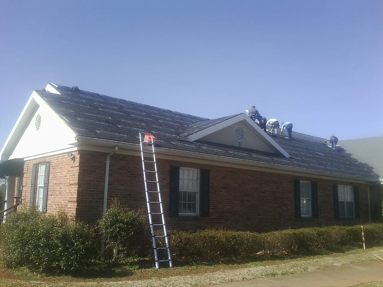 Crew tearing shingles off a brick ranch roof with a ladder set — Metal Roof Repair work by our crew in Lindenhurst