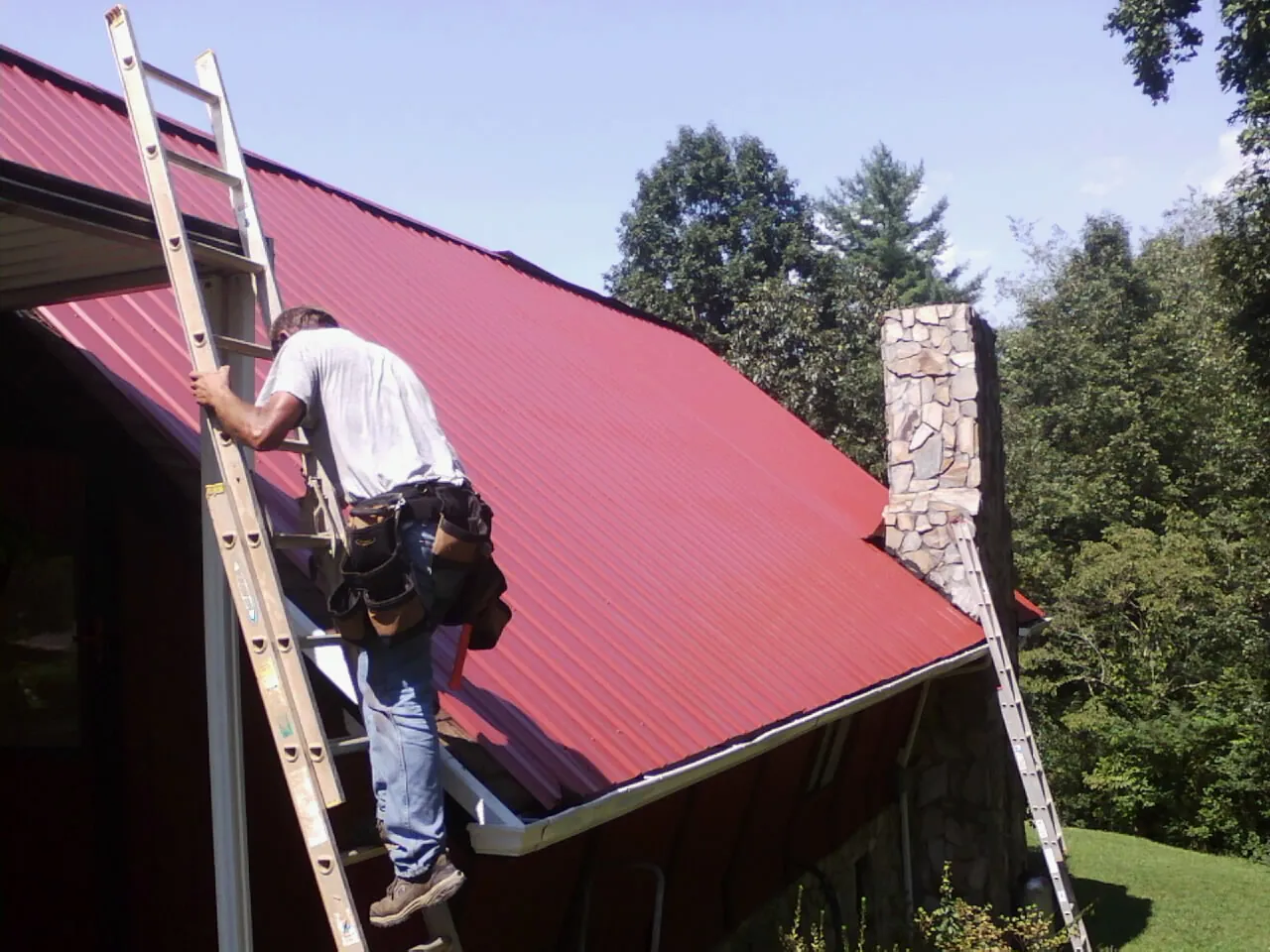Roofer on a ladder beside a red metal roof and stone chimney — Soffit & Fascia Repair by Roofing Brothers in Lindenhurst